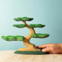 Close up of a hand holding a Bumbu handmade wooden Bonsai tree on a wooden worktop in front of a blue background