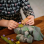 Close up of some children's hands playing with a Bumbu wooden deer and grass toy figures on a wooden table