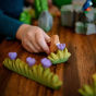 Close up of a hand holding a Bumbu wooden bunny animal figure on a wooden table next to a small grass toy with purple flowers