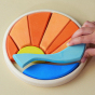 Circular wooden toy depicting the sun setting over the sea. A hand is in the photo showing the last piece being fitted to complete the scene.