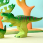 Wooden toy adult T-Rex in vibrant shades of green and purple. The T-Rex stands upright with its jaws open in a forward stance. The toy has painted teeth, claws, and eye detail. The toy is against a backdrop of wooden tree figures. In the foreground, there
