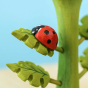A closer view of the Bumbu red wooden ladybird with painted black spots, stood on a green leaf which is attached to a green plant stalk, on a light blue background