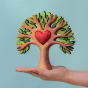 Bumbu Wooden Heart Tree. A beautifully crafted Wooden Tree with light brown branches, light and dark green leaves, with a removable wooden red heart in the centre. The tree is displayed on the palm of a hand  with a blue background