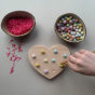 Child using tweezers to fill the indents on the Coach House Heart Fine Motor Board, a bowl of pompoms, and a bowl of pink rice. 