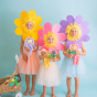 Three children wearing flower masks on their faces, holding Lavender Pickle, Buttercup Pip and Fuchsia Twinkle in their hands, along with paper flowers on a blue background