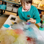 Child leaning over to blow coloured bubbles into a big pile on a white sheet of paper