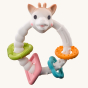 Sophie The Giraffe - So Pure Early Learning Gift Set. A multi textured, natural rubber teething ring with Sophie The Giraffe head, a green triangle ring, a pink diamond ring, and blue flower ring, and an orange circle ring all with different textures, on 
