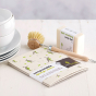 Pack of Ecoliving compostable sponge cleaning cloths laid out on a white table next to a wooden scrubbing brush, soap bar and a pile of white bowls