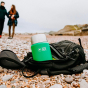 Close up of the green Elephant Box reusable insulated food flask in a black backpack on a beach