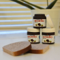 Three jars of Erzi Erzella Chocolate Spread Wooden Play Food stacked up, one with lid off and two slices of wooden toast in the foreground