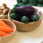 A natural woven basket holding a collection of Erzi Aubergine Eggplant Wooden Play Food surrounded by baskets of other toy veggies.