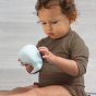 Young child with brown long sleeved dress and brown curly hair is playing with Hevea Squeeze'n'splash Whale Bath toy in Blizzard Blue. Looks very focussed, grey background 
