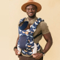Tula - A man carries a child on his chest wearing a Coast Whale Watch - Explore Baby Carrier. This is a front-on studio shot with a soft yellow background. The baby is facing the man's chest in the carrier.