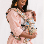 Tula - A woman carries a child on her chest wearing a Flower Walk - Explore Baby Carrier. This is a side-on studio shot with a pastel pink background.