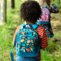 A child walks through the woods wearing the Frugi Little Adventurers Backpack - Fir Tree / Rainbow Leaves.