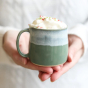 Glosters Pottery Sage Green Mug. A handmade stoneware mug by Glosters Pottery in Sage is a grey-green mug with fluid drippy white glaze, which looks like sea foam crashing against the beach, filled with a hot drink, whipped cream and sprinkles, being held