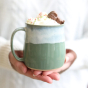 Glosters Pottery Sage Green Mug. A handmade stoneware mug by Glosters Pottery in Sage is a grey-green mug with fluid drippy white glaze, which looks like sea foam crashing against the beach, filled with a hot drink, whipped cream and a chocolate figure, b