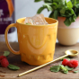 Glosters handmade Sandcastle Mug - Mustard Yellow. The mug sits on a wooden table, it is full of ice and has fruit and mint in front of it.