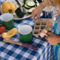 A child sorting various seeds and food items whilst holding onto one of the Grapat Wooden Sensory Play Tools. Different foods lay on the table which is covered in a blue and white chequred table cloth