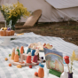 A picnic spread with cake, apples and a picnic blanket, with the Grapat Your Day Wooden Celebration Set on top. Hand painted wooden confetti coins, balls and figures including a photo holder, vase, Nin and candle in rainbow colours.