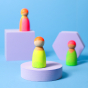 Grimm's 3 Friends Wooden Peg People in a Neon Mix of colours in pink, orange yellow and green displayed on a white stand with blue background.