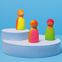 Grimm's 3 Friends Wooden Peg People in a Neon Mix of colours in pink, orange yellow and green displayed on a white stand with blue background.