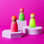 Grimm's 3 Friends Wooden Peg People in a Neon Mix of colours in pink, orange yellow and green displayed on a white stand with bright pink background.