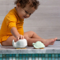A small child in a long sleeved yellow playsuit, plays with the Hevea Squeeze'n'splash Bath toys - Whale & Turtle - Frosty White - Sage, at the side of a pool. Grey background, varying shade mini blue / green tiles at side of pool.
