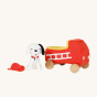 Front view of Olli Ella Holdie Dog-go Chief sat next to a red felt fire truck with wooden wheels, and red fire fighter helmet on a cream background