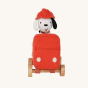 Front view of Olli Ella Holdie Dog-go Chief in a red felt fire truck with wooden wheels, wearing a red fire fighter helmet on a cream background