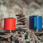 The Klean Kanteen Camping Mug in Red and Dark Denim, in front of a small rock formation. The Dark Denim mug shows the Klean Kanteen Logo on the handle.
