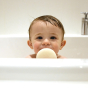 A baby sits in a white bath, peering over the edge towards the camera with Kokoso Natural Baby Konjac Sponge in their hands, just in front of their mouth.