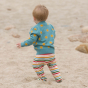 Young child walking on some sand in rainbow striped trousers and a blue LGR sunshine cardigan