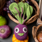 Close up of the Myum Emily beetroot plastic free cotton soft toy leaned against a wicker bowl of grapes