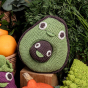 Myum avocado crochet soft toy on a wooden block next to some green and orange vegetables