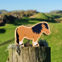 Close up of the Lanka Kade shetland pony toy figure stood on a wooden post in front of some green fields