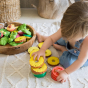 A small child playing with Oli & Carol Ananas the Pineapple natural rubber teether and other Oli & Carol items, on a natural rug, a wooden bowl with lots of Oli & Carol fruit and veg teether sits to the side