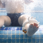 Picture of a baby sat in a pool, with the Oli and Carol Sharon the Crab rubber bath toy around their ankle.