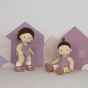 Two Olli Ella Bitsy Dinkum Dolls with both with long brown hair, blue eyes and white skin, wearing a purple knitted romper style outfit. On has their hair down and one has it up. They are pictured against a lilac coloured background with cut-out house and
