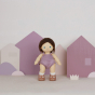 Olli Ella Bitsy Dinkum Doll with long brown hair, blue eyes and white skin, wearing a purple knitted romper style outfit. Pictured on a lilac coloured background with cut-out house and grass shapes