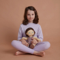 A child sitting cross legged holding the Olli Ella Bitsy Dinkum Doll on their lap. The child is looking at the camera. Both child and doll are wearing purple clothing. Pictured on a plain terracotta coloured background.