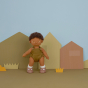 An Olli Ella Button Dinkum Doll pictured against a multi coloured background with cut out shapes of houses and grass. The doll is stood up with their arms down by their side