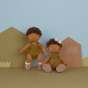 Two Olli Ella Button Dinkum Doll pictured against a multi coloured background with cut out shapes of houses and grass. One doll is stood up with their arms down by their side, the other is sat down next to them wearing a white coloured bow in their hair 