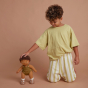 A curly haired child kneeling next to an Olli Ella Button Dinkum Doll. The doll is also stood up next to them, the child has their hand placed on the dolls head. They are pictured in front of a dark terracotta coloured background 