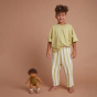 A curly haired child standing next to an Olli Ella Button Dinkum Doll. The doll is also stood up next to them. They are pictured in front of a dark terracotta coloured background 