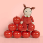 Olli ella annie apple dinky doll toy sat on a pile of red apples in front of a pink background