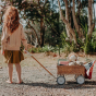 Girl stood on some gravel pulling the Olli Ella rattan wonder wagon in the natural colour filled with toys and books