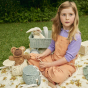 Girl sat on a floral blanket on some grass playing with a cozy dozy doll and the olli ella eco-friendly rattan chari basket