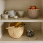 Olli Ella large rattan blossom basket, filled with bananas on a kitchen cupboard shelf next to a teapot and a jar of herbs