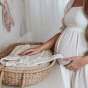 Pregnant woman in a white dress, placing her hand on top of some knitted baby clothing in an Olli Ella rattan basket with a luxe liner cushion
